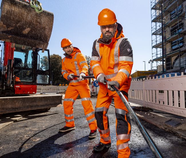 Straßenbauer verlegen einen Kabel und tragen Warnschutzkleidung in orange.